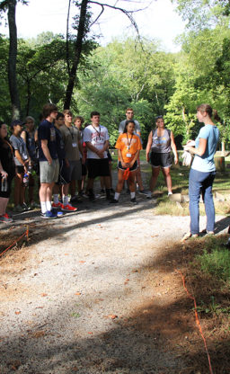 A woman in the foreground speaks to a large gathering of students standing on the path that is under construction.
