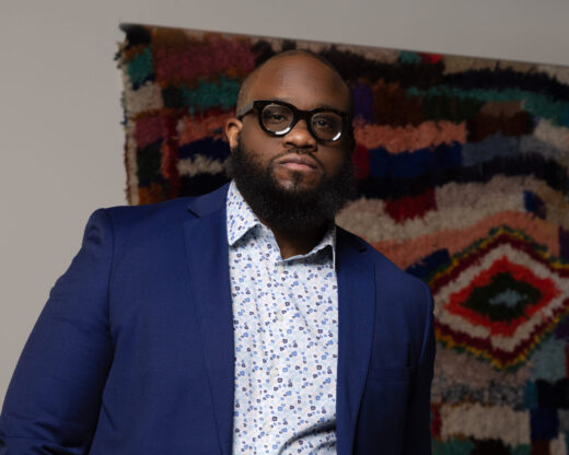 Joseph Cole, a Black man wearing a patterned button-up with a blue blazer and glasses, stands in front of a colorful wall piece.