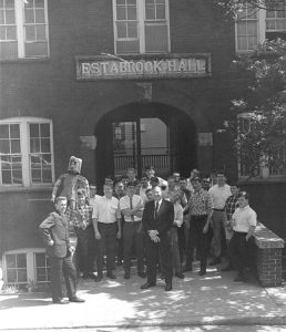 An old black and white photo shows a group of man standing in front of the brick Estabrook Hall