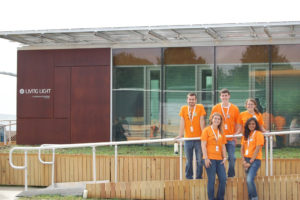 The team stands in front of the house wearing orange shirts.