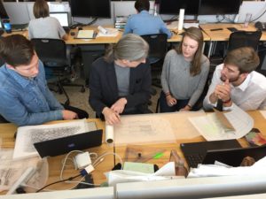 A faculty member sits at a table discussing architectural drawings with two students.