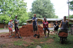 A group of students with shovels stand around a large pile of dirt while another male student pushes dirt in a wheelbarrow.