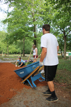 A male student dumps a wheelbarrow load of dirt onto the path where two female students are working.