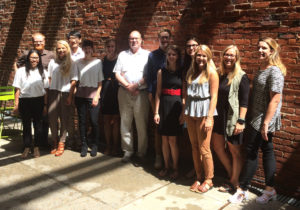 A group of 13 students and faculty stand against a brick wall for a group photo