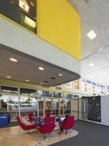An open two-story room within the Art + Architecture building with students sitting in red chairs in the foreground.