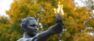 A bronze statue holding a flame against a backdrop of fall leaves.