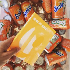 A hand holding a "Nine by Nine" book over a cooler filled with Sunkist soda.