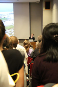 A woman is visible giving a lecture in the background with an audience in the foreground.