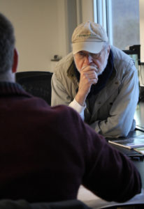 A gray-haired man sits and thinks while looking down at a document on the table.
