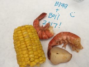 Two cooked shrimp and a corn cob on a table at the Brag + Boil event.