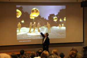 A man giving a lecture in front of an audience gestures to an image on the screen.