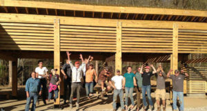 A group of students and faculty celebrate in front of the wooden shelter they have just built.