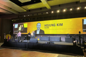 The stage at the American Society of Interior Designers' conference. On the screen, a yellow background features an image of Hojung Kim with his name and title 'Assistant Professor, University of Tennessee, Knoxville'. Hojung poses with a woman for a photo.