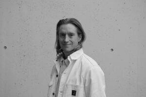 Chachi Mathis, a white male with shoulder-length hair. He is wearing a white jacket and button up shirt.