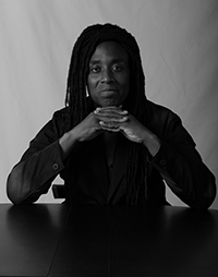 A black-and-white portrait of Anthony Titus with long braided hair, wearing a dark jacket, seated at a table with their hands clasped together in front of them. The subject, a Black male, looks directly into the camera with a composed expression.