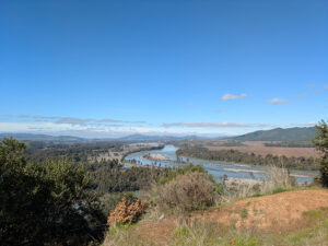 This image shows a beautiful landscape with a river winding through a valley surrounded by lush greenery and mountains in the distance. The terrain and features suggest it could be from a region in Chile known for its natural beauty, in the Central Valley, which includes areas like the Maule or Bio-Bio regions. These areas are known for their rivers, rolling hills, and agricultural landscapes.