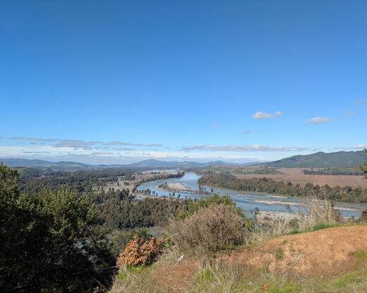 This image shows a beautiful landscape with a river winding through a valley surrounded by lush greenery and mountains in the distance. The terrain and features suggest it could be from a region in Chile known for its natural beauty, in the Central Valley, which includes areas like the Maule or Bio-Bio regions. These areas are known for their rivers, rolling hills, and agricultural landscapes.