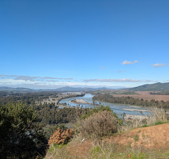 This image shows a beautiful landscape with a river winding through a valley surrounded by lush greenery and mountains in the distance. The terrain and features suggest it could be from a region in Chile known for its natural beauty, in the Central Valley, which includes areas like the Maule or Bio-Bio regions. These areas are known for their rivers, rolling hills, and agricultural landscapes.