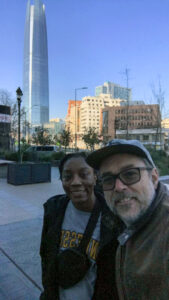 Ebone, on the left, and Germán, on the right, smile for a selfie in Santiago, Chile, with the towering Gran Torre Santiago skyscraper and city buildings visible in the background on a clear day.