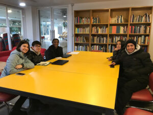 Five students, Camila, Jose, Ebone, Carla, and Camilo, sit together at a yellow table in a bright, modern library at the University of Talca in Chile. They are wearing warm clothing, smiling, and ready to work on their studies, with bookshelves filled with colorful books in the background.