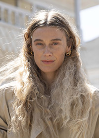 A color headshot of Lucy McRae with long, wavy, blonde hair and gold hoop earrings, standing outdoors in soft natural light. She is a white woman are wearing a light tan jacket and have a calm, serene expression.