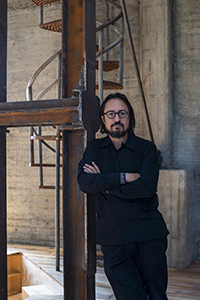 A portrait of Javier Sánchez with shoulder-length dark hair and glasses, standing with arms crossed in front of a metal staircase within an industrial building. The Mexican male is dressed in black and leans against a large rusted metal structure, looking directly at the camera.