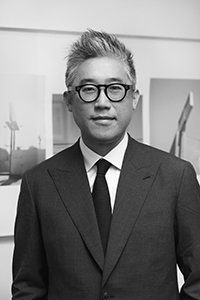 A black and white portrait of Mark Lee wearing round glasses, a white button up with a tie and dark colored jacket. The Asian male looks directly into the camera with a composed expression.