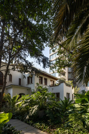 A photograph of the Juan de la Barrera residential complex, showing a beautifully landscaped garden with dense greenery surrounding a historic, two-story building. The building has a light-colored façade and traditional architectural details, with a modern glass tower visible in the background. Photo by Rafael Gamo.