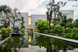 A view of the MMAC Juan Soriano Museum, featuring a modern, minimalistic concrete building. The museum is surrounded by a tranquil water feature and lush, tropical vegetation, with a large sculpture visible in the foreground. Photo by Rafael Gamo.