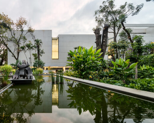 A view of the MMAC Juan Soriano Museum, featuring a modern, minimalistic concrete building. The museum is surrounded by a tranquil water feature and lush, tropical vegetation, with a large sculpture visible in the foreground. Photo by Rafael Gamo.