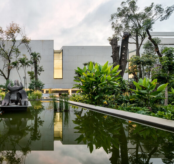 A view of the MMAC Juan Soriano Museum, featuring a modern, minimalistic concrete building. The museum is surrounded by a tranquil water feature and lush, tropical vegetation, with a large sculpture visible in the foreground. Photo by Rafael Gamo.