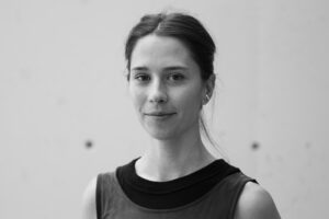 Black and white headshot of Lauren Buntemeyer, a white woman, with her dark hair pulled back, wearing a sleeveless top, and offering a subtle smile, standing against a plain light-colored background.