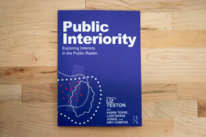 Cover of the book titled 'Public Interiority: Exploring Interiors in the Public Realm' edited by Liz Teston, with Karin Tehve, Ladi'Sasha Jones, and Amy Campos. The cover is blue with white text and abstract graphic elements in light blue, white, and red. The book is resting on a wooden surface.