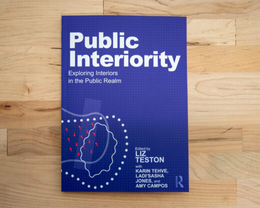 Cover of the book titled 'Public Interiority: Exploring Interiors in the Public Realm' edited by Liz Teston, with Karin Tehve, Ladi'Sasha Jones, and Amy Campos. The cover is blue with white text and abstract graphic elements in light blue, white, and red. The book is resting on a wooden surface.