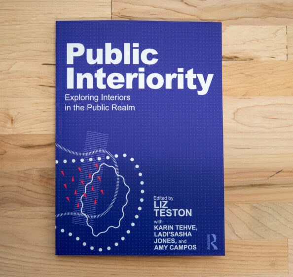 Cover of the book titled 'Public Interiority: Exploring Interiors in the Public Realm' edited by Liz Teston, with Karin Tehve, Ladi'Sasha Jones, and Amy Campos. The cover is blue with white text and abstract graphic elements in light blue, white, and red. The book is resting on a wooden surface.