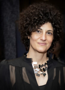 A color photo of Aroussiak Gabrielian, an Armenian-born woman with dark, short curly hair. She is wearing a black blouse with a black tank top underneath and a chunky metal necklace.