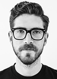 A black and white headshot of Jesse Reed, a man with with short, neatly styled hair and a full beard and mustache. He wearing rectangular, thick-framed glasses and a plain black T-shirt with a pocket on the chest.