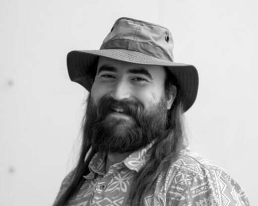 Black-and-white headshot of Thomas Cox, a man with a long beard and long hair wearing a patterned shirt and a bucket hat, smiling.