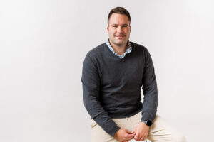 Professional headshot of Joey Staats, a white male, wearing a gray sweater over a checkered collared shirt and beige pants, seated against a plain white background with a relaxed, approachable expression.