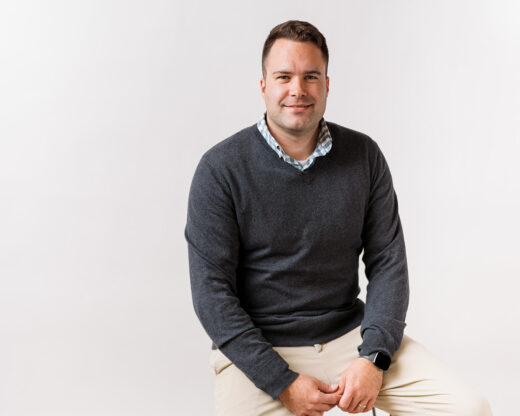 Professional headshot of Joey Staats, a white male, wearing a gray sweater over a checkered collared shirt and beige pants, seated against a plain white background with a relaxed, approachable expression.