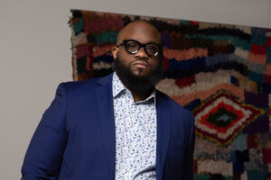 Joseph Cole, a Black man wearing a patterned button-up with a blue blazer and glasses, stands in front of a colorful wall piece.