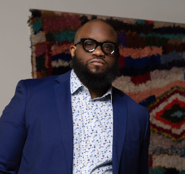 Joseph Cole, a Black man wearing a patterned button-up with a blue blazer and glasses, stands in front of a colorful wall piece.