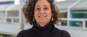 Headshot of Avigail Sachs smiling, with short curly brown hair, wearing a dark sweater and earrings, standing indoors with a softly blurred modern architectural background.