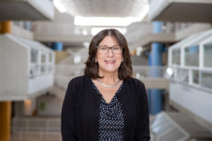 Pamela Cannella Treacy, a woman with shoulder-length brown hair and glasses, smiles while standing in the modern, multi-level atrium of the Art + Architecture Building at the University of Tennessee, Knoxville. She is wearing a black cardigan over a black and white polka dot blouse, with a silver necklace.