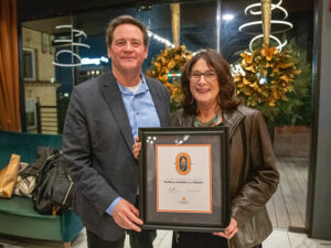 Pamela Cannella Treacy, wearing a black leather jacket and a teal necklace, stands next to Dean Jason Young, who is dressed in a dark suit with a blue button-down shirt. They are both smiling while holding a framed certificate for the Spirit of a Volunteer award. The background features festive golden wreaths and a warmly lit venue.