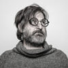 Smiljan Radić, a male with medium, shaggy hair covering round glasses looks off in the upper right corner. He is wearing a loose turtle-neck shirt in an almost greyscale image.