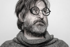 Smiljan Radić, a male with medium, shaggy hair covering round glasses looks off in the upper right corner. He is wearing a loose turtle-neck shirt in an almost greyscale image.