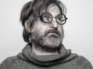 Smiljan Radić, a male with medium, shaggy hair covering round glasses looks off in the upper right corner. He is wearing a loose turtle-neck shirt in an almost greyscale image.