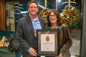 Pamela Cannella Treacy, wearing a black leather jacket and a teal necklace, stands next to Dean Jason Young, who is dressed in a dark suit with a blue button-down shirt. They are both smiling while holding a framed certificate for the Spirit of a Volunteer award. The background features festive golden wreaths and a warmly lit venue.