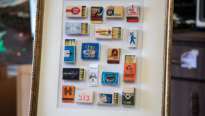 Framed artwork composed of vintage matchbooks arranged in a grid, each with different logos and illustrations, displayed on an easel.